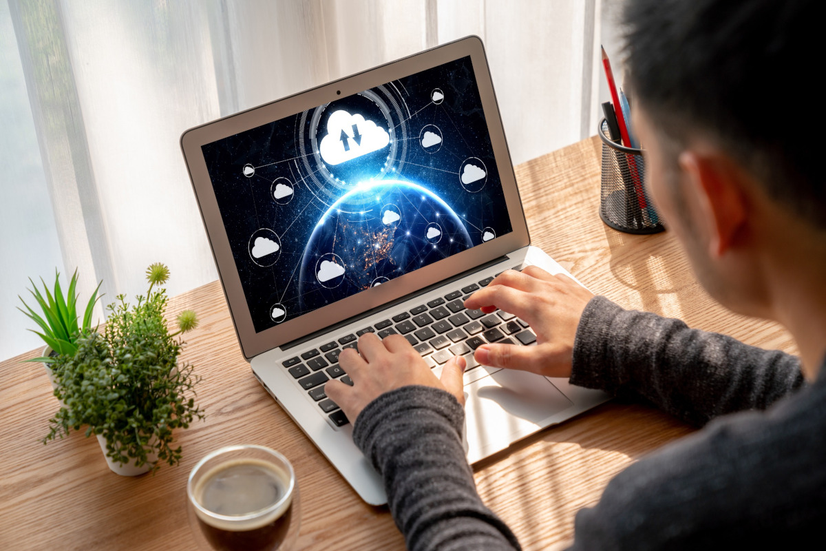Person typing on a laptop displaying a digital cloud network over a globe. Wooden desk, plant, coffee, and pencils in the background